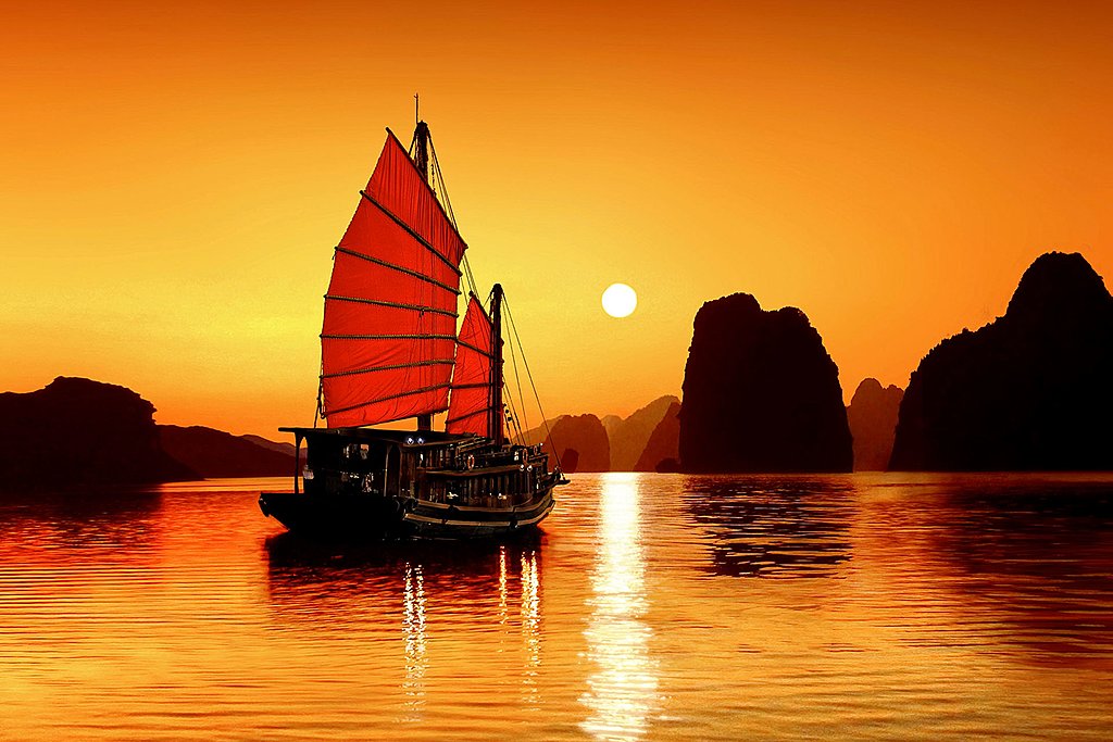 A traditional Vietnamese boat sailing through the limestone karsts of Ha Long Bay at sunset.