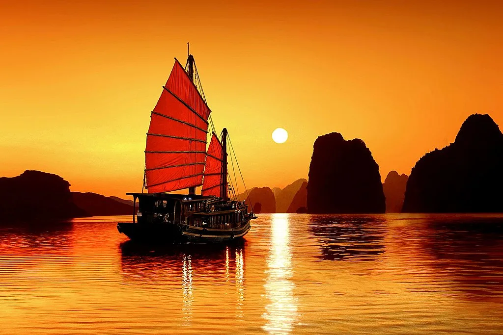 A traditional Vietnamese boat sailing through the limestone karsts of Ha Long Bay at sunset.